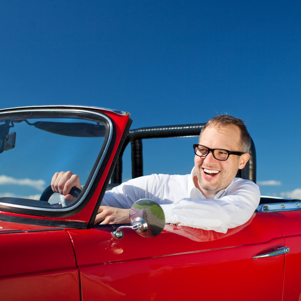 Lachender Brillenträger sitzt in einem roten Cabrio und schaut nach vorne.