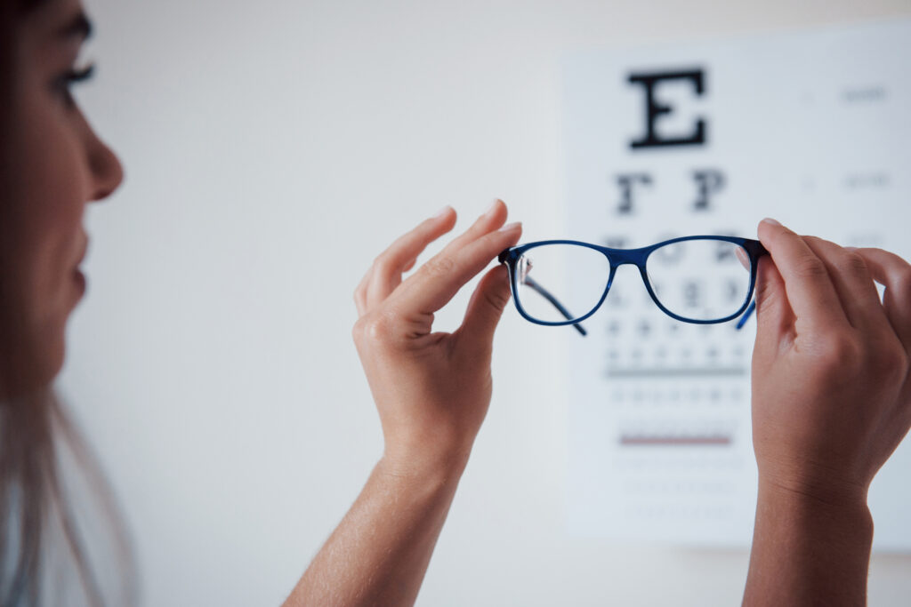 Frau hält eine Brille fest. Im Hintergrund erkennt man in der Unschärfe eine Sehtesttafel.