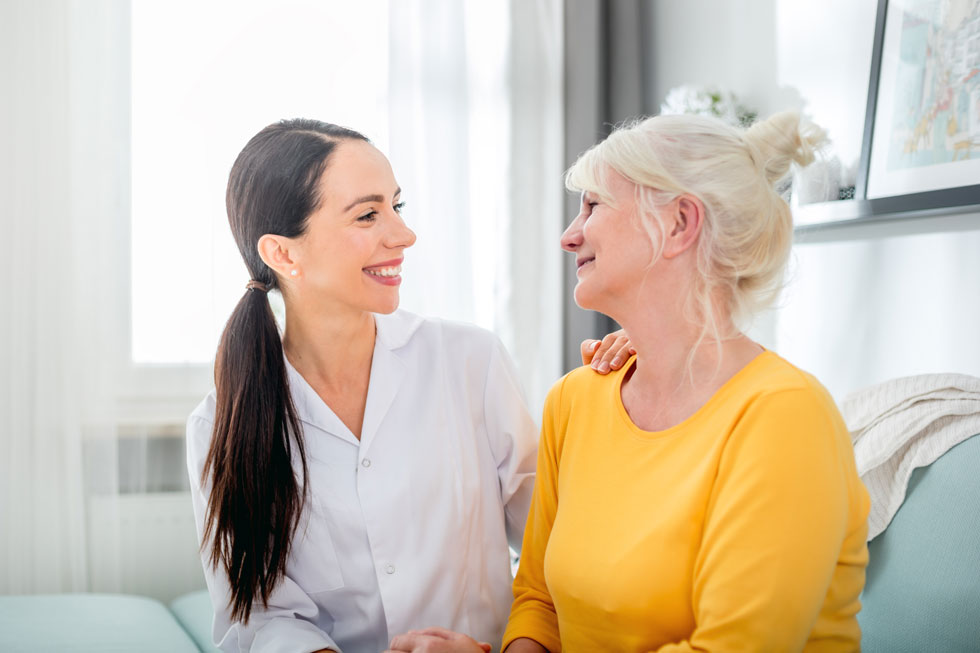 Junge Frau lächelt eine ältere Frau an und legt ihre Hand auf die Schulter Die beiden sitzen auf einem Sofa in einem Wohnzimmer.
