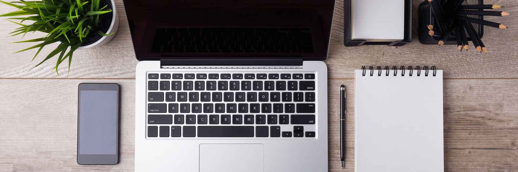 Vogelperspektive eines Schreibtisches, auf dem sich ein Laptop, ein Handy, Stifte und ein Notizblock befinden. Drer Betrachter schaut von oben auf den Schreibtisch.