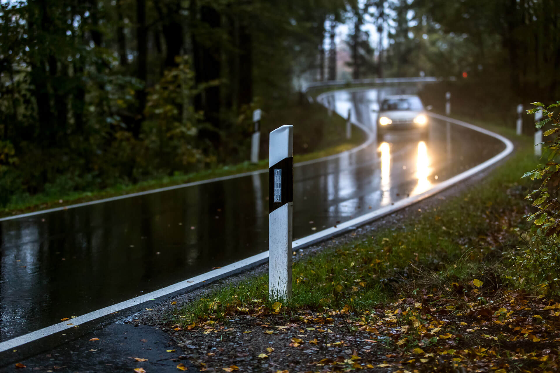 Blick auf eine verregnete Straße, auf der sich ein Auto nähert. Im Vordergrund ein Fahrbahnmarkierung