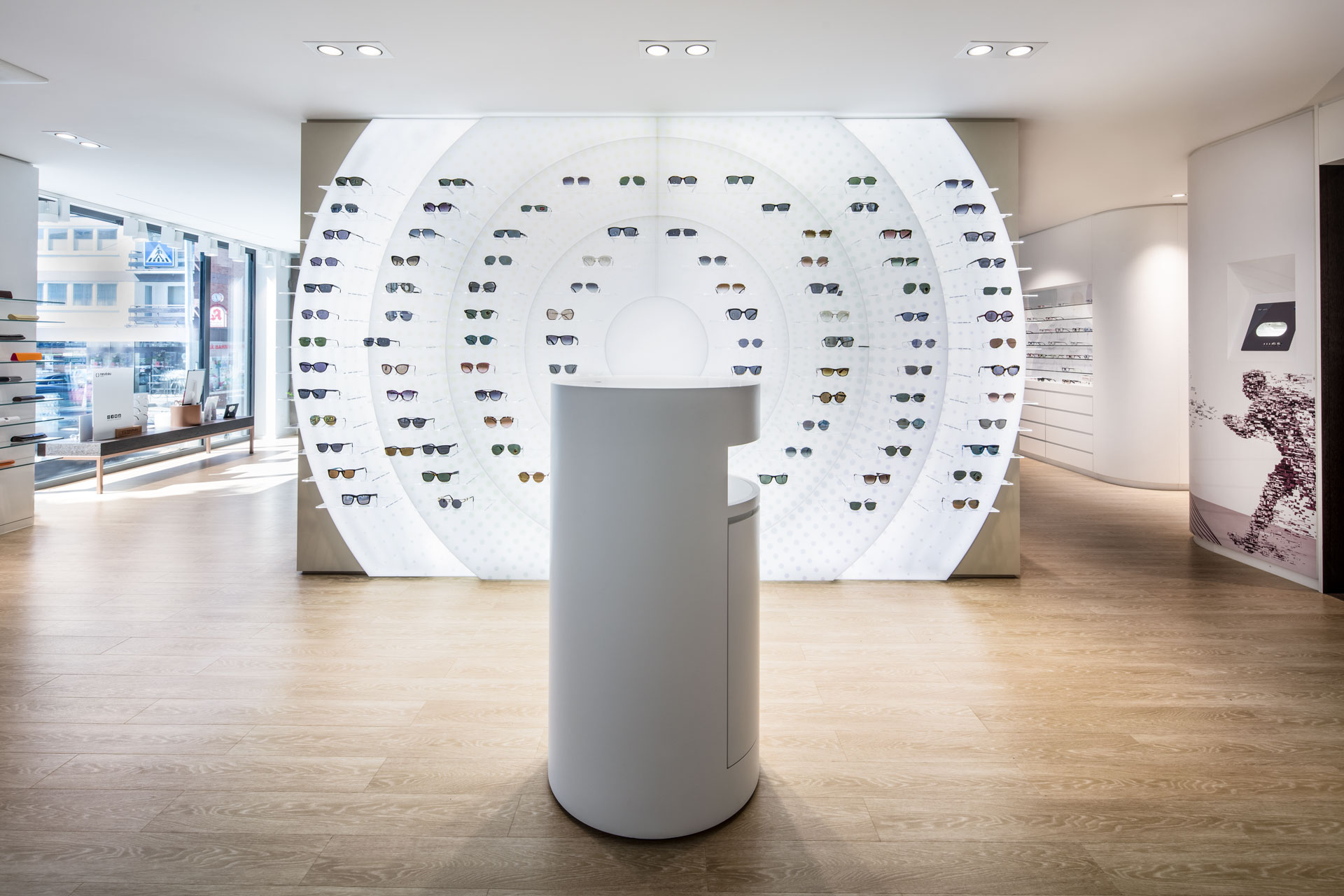 Frontaler Blick auf die beleuchtete Sonnenbrillen-Wand im Eingangsbereich Wittlich, an der eine große Auswahl an Sonnenbrillen präsentiert wird.