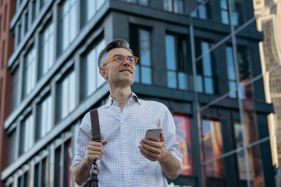 Älterer Mann mit orangenen Brille und Smartphone in der Hand mit Blick nach rechts, im Hintergrund ein hohes Gebäude