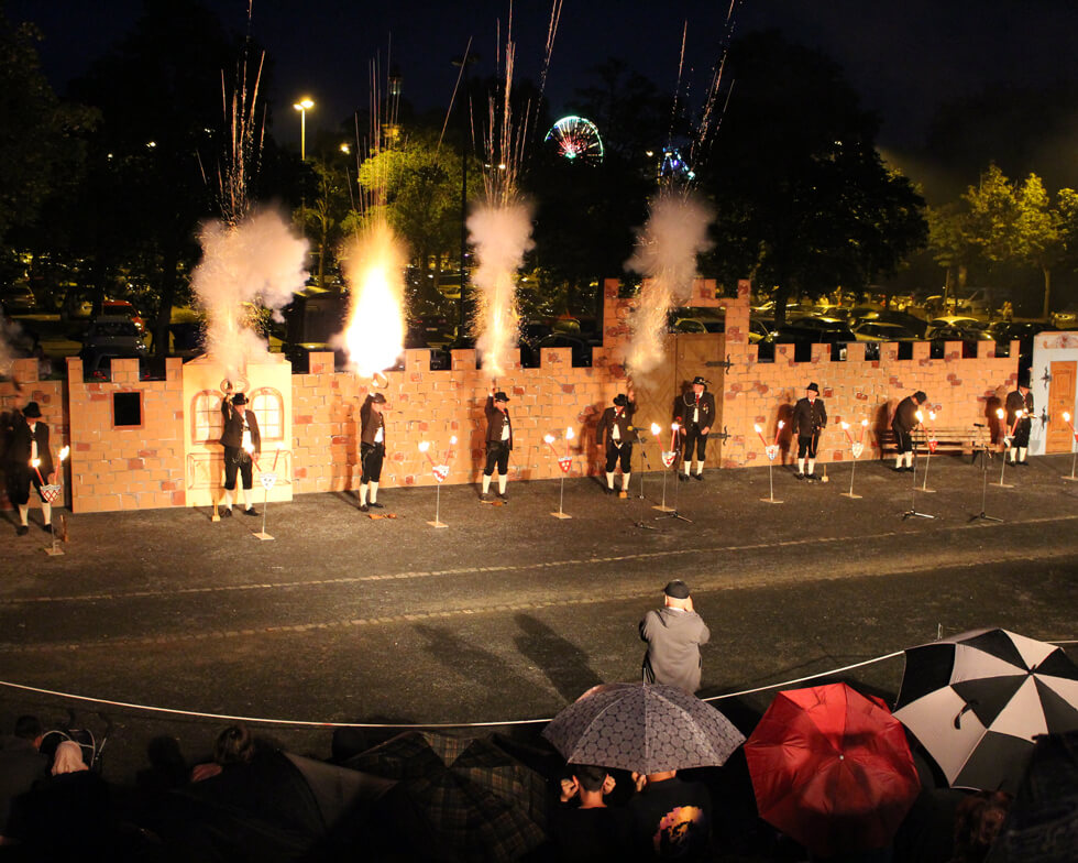 Mehrere Böllerschützen beim Säubrenner-Schauspiel, die in die Luft feuern. Im Hintergrund Rommelsbachparkplatz und am Horizont ein Riesenrad