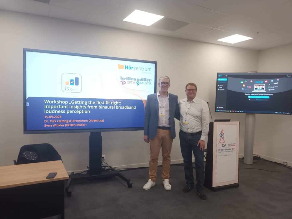Sven Winkler und Dr. Dirk Oetting in einem Tagungsraum auf dem WorldCongress of Audiology, im Hintergrund 2 große Fernseher mit einem Standbild des Workshops "Getting the first-tip right"