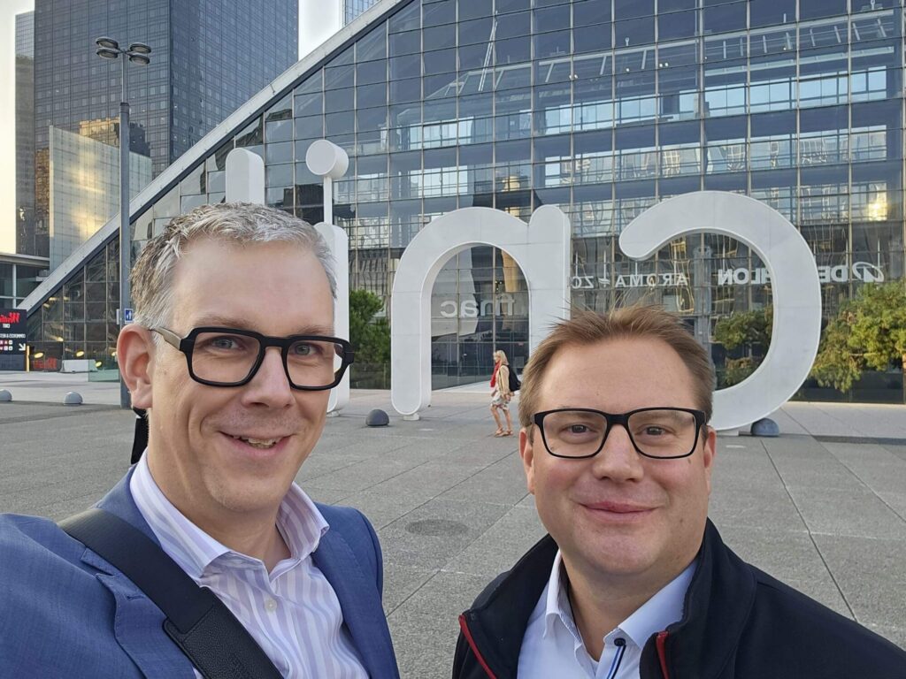 Sven Winkler und Dr. Dirk Oetting vor einem Glasgebäude des Hörkongress in Paris